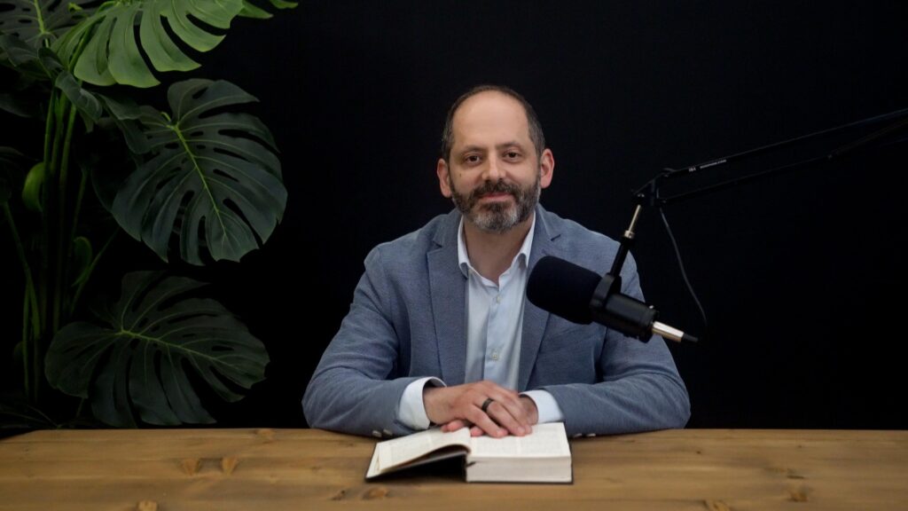 Man sits behind a wooden table with an open Bible ready to speak into a professional podcasting microphone | The Wisdom Journey by Endure International
