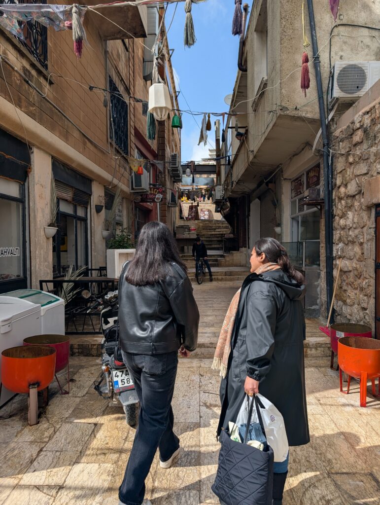 Two women wearing all black outfits face an alleyway between stone buildings. Lanterns with tassels are strung between the buildings above them.
