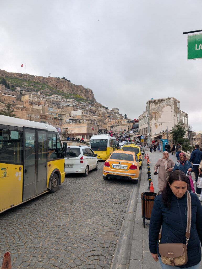 Yellow buses and taxis drive on an old cobblestone street through a historic central asian city built into a hillside. Cloud cover is gray and muted.