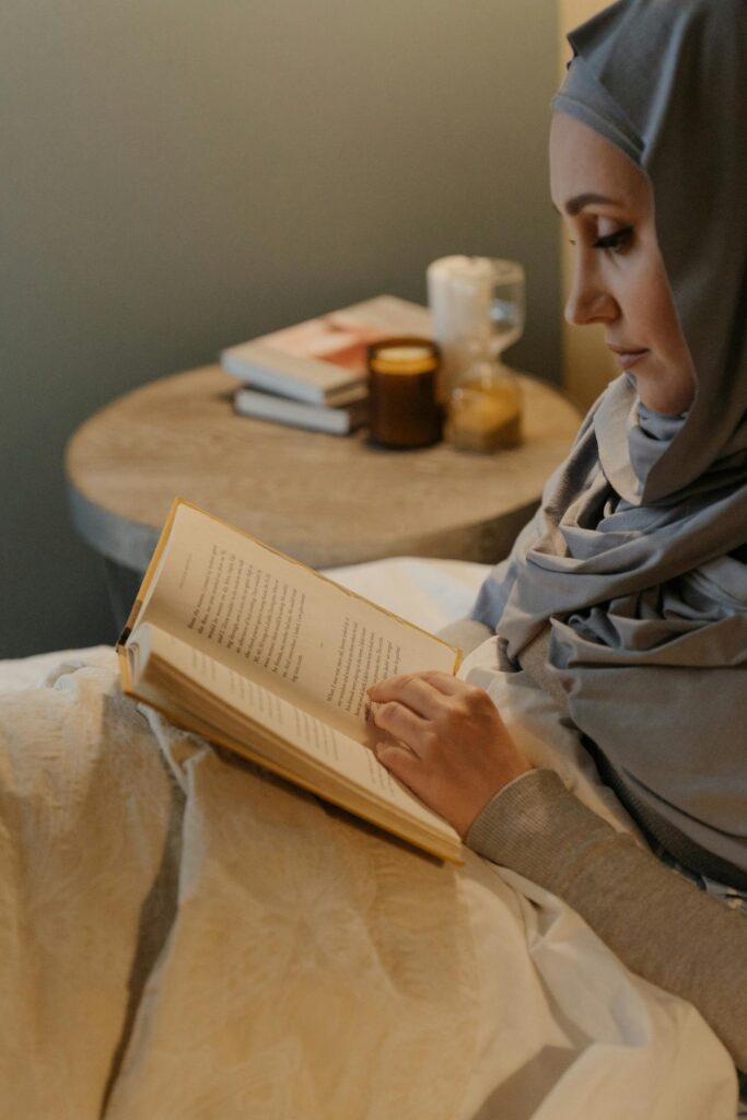 Woman wearing a head covering sits next to a small side table and reads a book