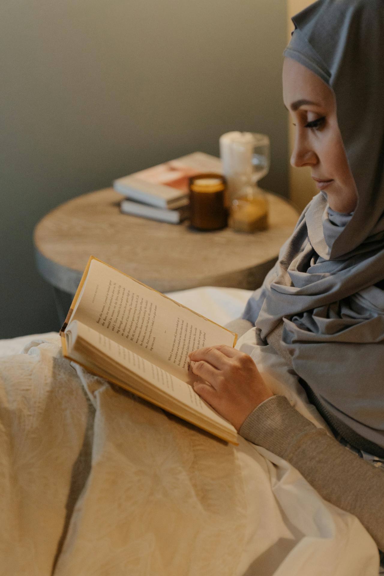Woman wearing a head covering sits next to a small side table and reads a book